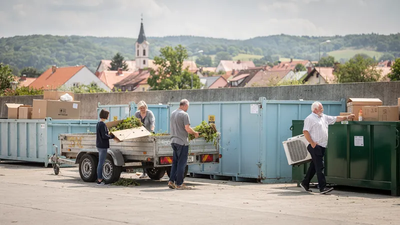 Menschen geben an einem tschechischen Sammelhof Grünschnitt und ein altes Klimagerät ab, Kleinstadt mit Kirchturm im Hintergrund.
